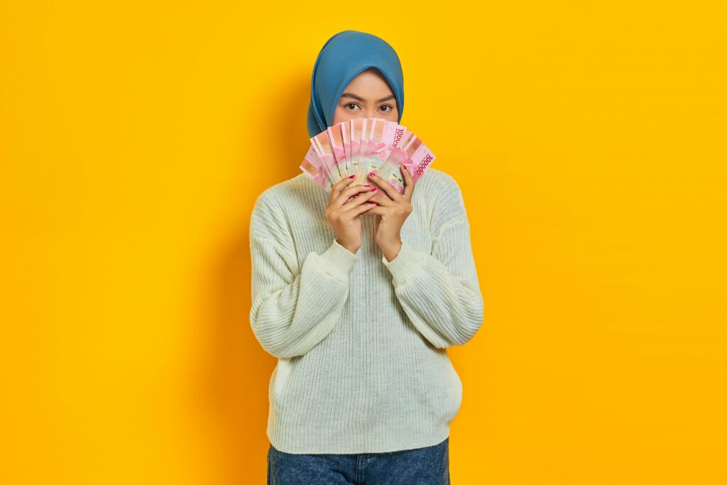 Portrait of a woman in hijab holding Rupiah notes against a yellow background, symbolizing wealth.